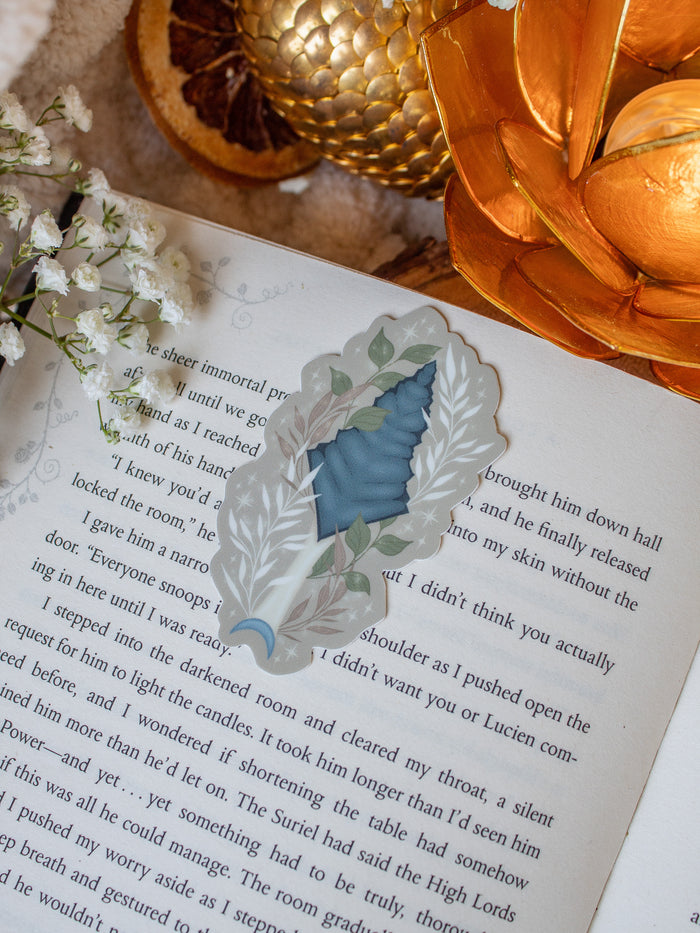 This bookish When The Moon Hatched dragonscale dagger sticker is inspired by the Moonfall series by Sarah A Parker. The image shows the sticker on an open book, with the book placed on a fuzzy neutral colored blanket, a faux golden dragon egg, orange slices, and an orange lotus shaped tea light holder all surrounded by little white flowers