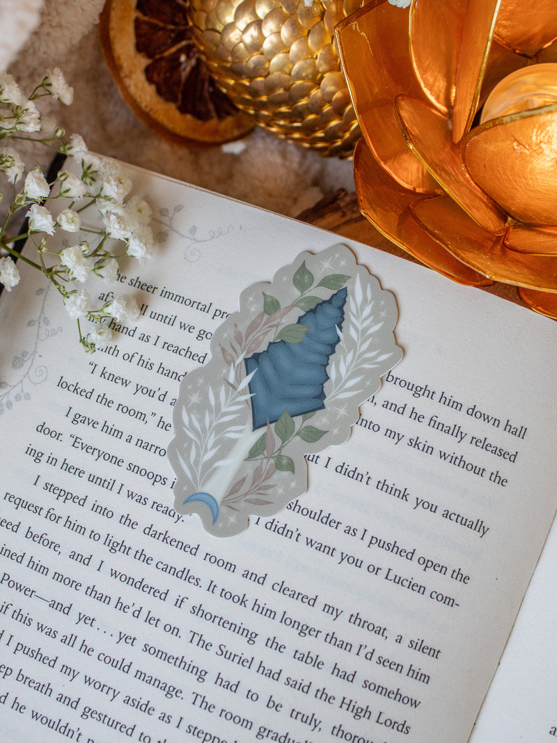 This bookish When The Moon Hatched dragonscale dagger sticker is inspired by the Moonfall series by Sarah A Parker. The image shows the sticker on an open book, with the book placed on a fuzzy neutral colored blanket, a faux golden dragon egg, orange slices, and an orange lotus shaped tea light holder all surrounded by little white flowers