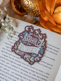 This When the Moon Hatched Sticker reads "So many assholes, so few fingers to count them all" in cream text on a blood red background with green and slate blue foliage and little stars, magic bubbles, and dragons flying around. 