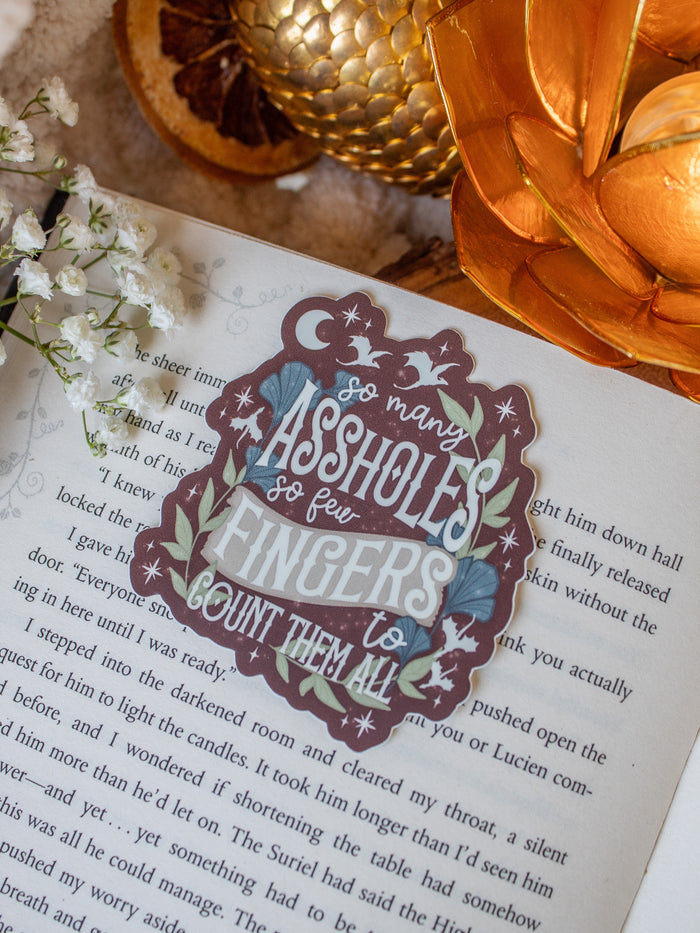This When the Moon Hatched Sticker reads "So many assholes, so few fingers to count them all" in cream text on a blood red background with green and slate blue foliage and little stars, magic bubbles, and dragons flying around. 