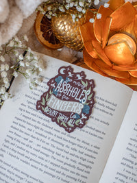 This When the Moon Hatched Sticker reads "So many assholes, so few fingers to count them all" in cream text on a blood red background with green and slate blue foliage and little stars, magic bubbles, and dragons flying around. 