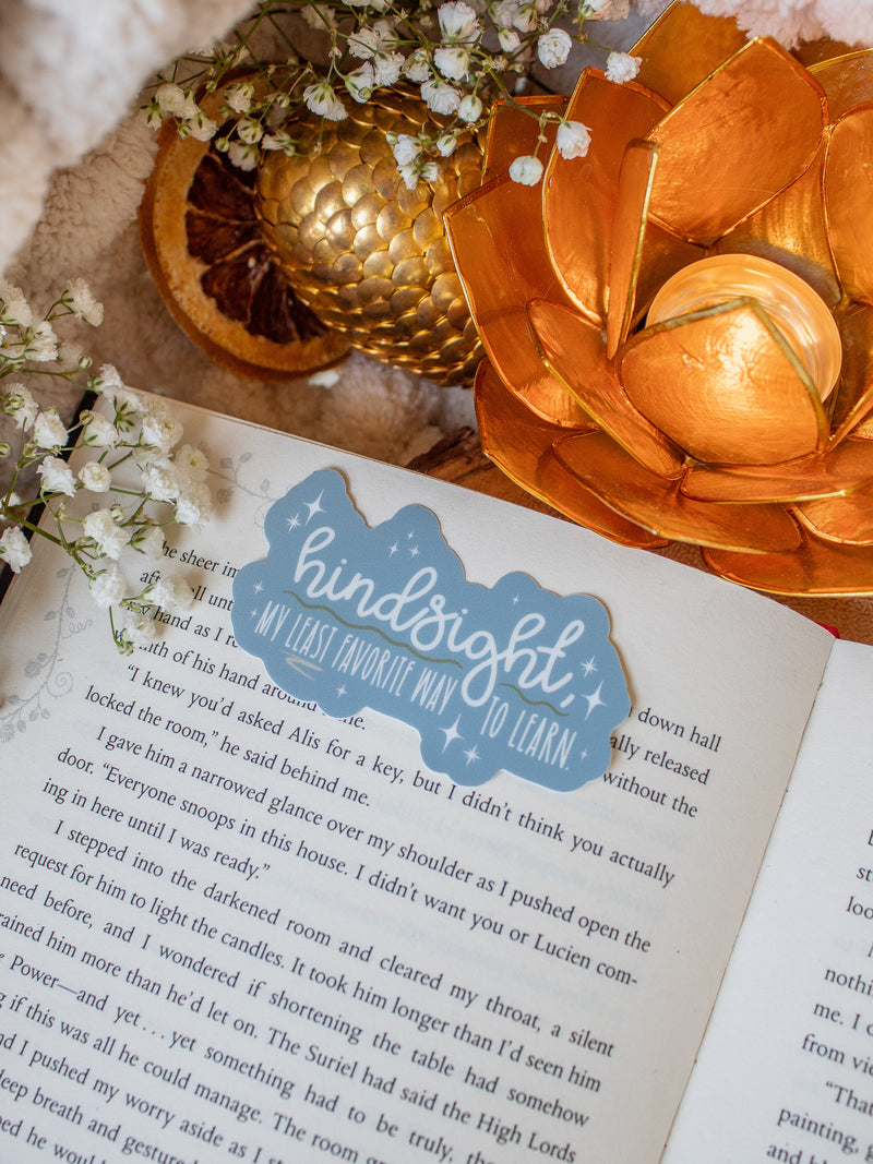 This When the Moon Hatched Sticker from the Moonfall Series by Sarah A Parker is a light blue with the words "Hindsight, my least favorite way to learn" on it in white with green and taupe annotation style accents around them. 