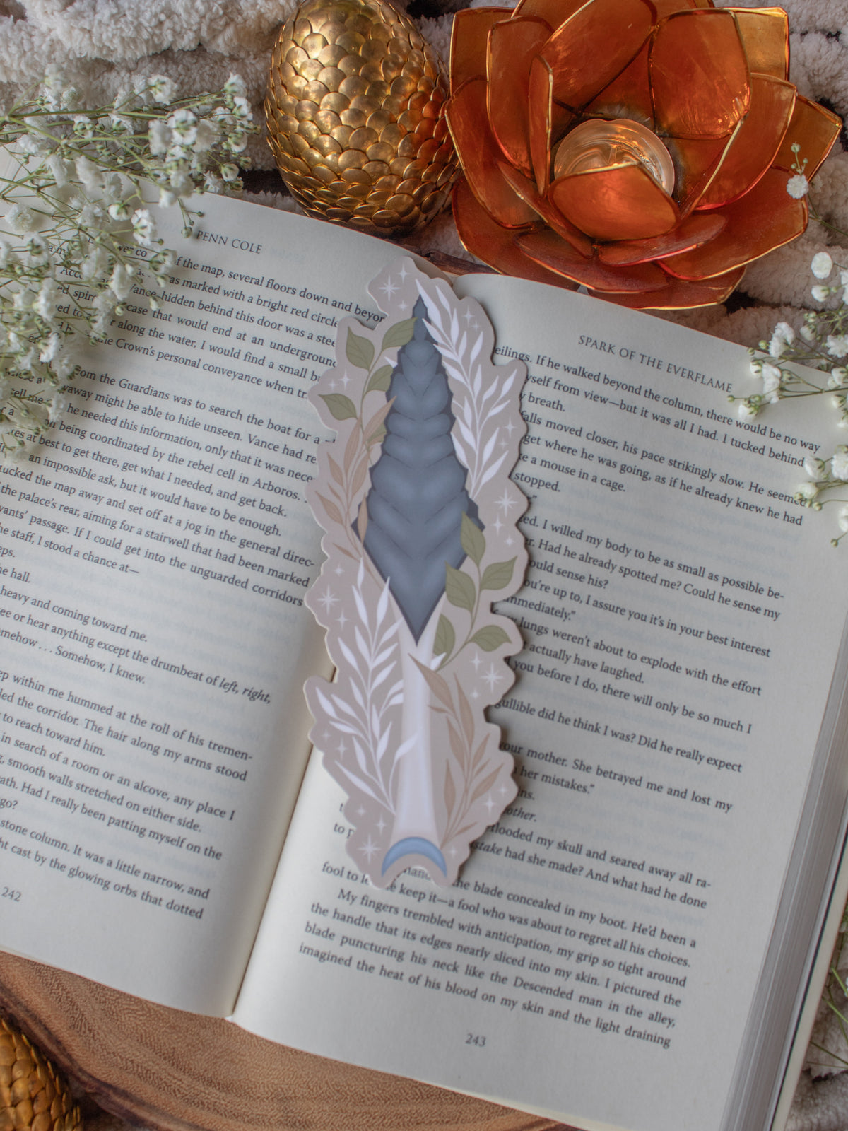 This bookish When The Moon Hatched dragonscale dagger die-cut bookmark is inspired by the Moonfall series by Sarah A Parker. The image shows the bookmark on an open book, with the book placed on a fuzzy neutral colored blanket, a faux golden dragon egg, orange slices, and an orange lotus shaped tea light holder all surrounded by little white flowers