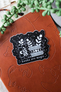 This "Books Are Magic" waterproof vinyl sticker has a black background with a white design and text. The design is a small bookshelf with 6 books. The 3 on the left are vertical with a moon, serpent, and stars on them. The three on the right are stacked horizontally with the top and bottom one showing the pages and the middle ones spine has a moon phase image. The books are surrounded by foliage and little stars.