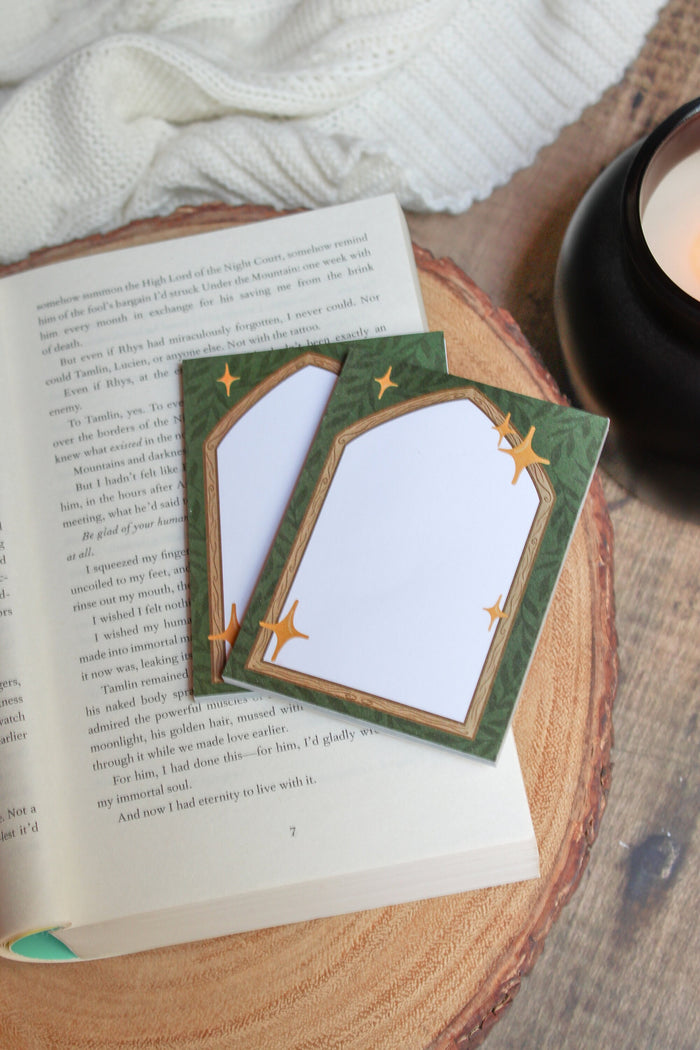 These Fantasy Bookish Archway Post-It Notepads have a dark green background with lil leaves on it. In front their is a wooden archway that comes to a point at the top. The center of the archway is solid write for the writing area. 