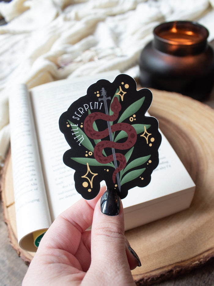 This Little Serpent sticker from The Serpent and The Wings of Night from the Crowns of Nyaxia Series by Carissa Broadbent is officially licensed. It features a maroon snake wrapped around a steel sword in front of green foliage on a black background with the text "Little Serpent" around the body of the snake. 