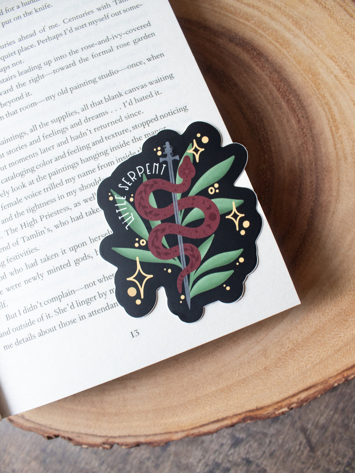 This Little Serpent sticker from The Serpent and The Wings of Night from the Crowns of Nyaxia Series by Carissa Broadbent is officially licensed. It features a maroon snake wrapped around a steel sword in front of green foliage on a black background with the text "Little Serpent" around the body of the snake. 