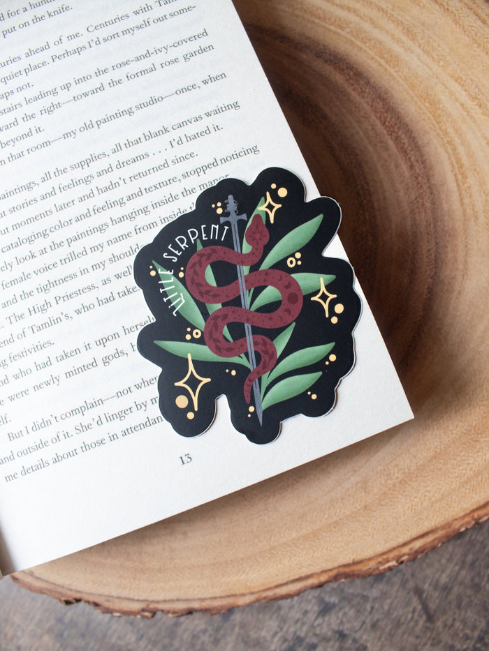This Little Serpent sticker from The Serpent and The Wings of Night from the Crowns of Nyaxia Series by Carissa Broadbent is officially licensed. It features a maroon snake wrapped around a steel sword in front of green foliage on a black background with the text "Little Serpent" around the body of the snake. 