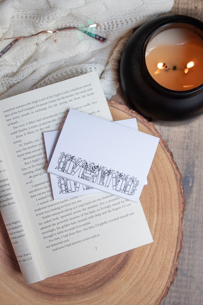 These bookish magical forest bookshelf post-it notepads are perfect to jot down all your bookish thoughts and ideas. They are 3"x4", have a white background with a black bookshelf along the bottom. The bookshelf have foliage peeking out from behind them. 