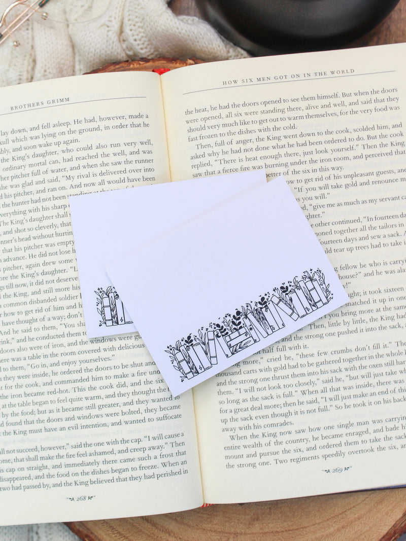 These bookish magical forest bookshelf post-it notepads are perfect to jot down all your bookish thoughts and ideas. They are 3"x4", have a white background with a black bookshelf along the bottom. The bookshelf have foliage peeking out from behind them. 