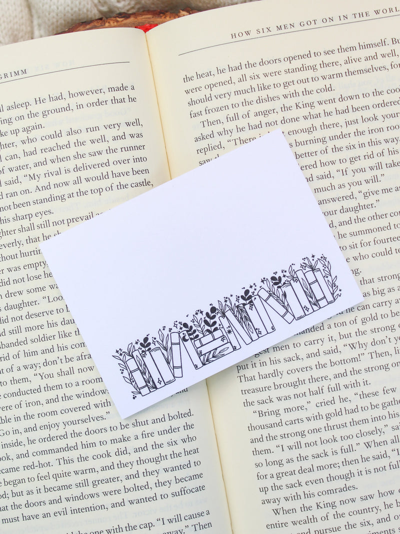 These bookish magical forest bookshelf post-it notepads are perfect to jot down all your bookish thoughts and ideas. They are 3"x4", have a white background with a black bookshelf along the bottom. The bookshelf have foliage peeking out from behind them. 
