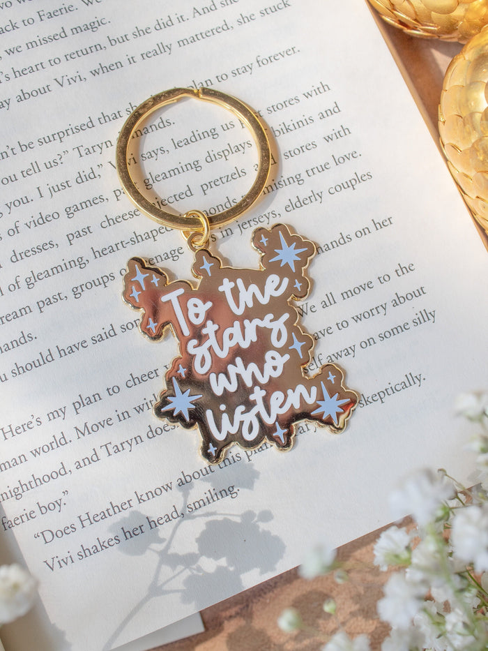 This officially licensed "To The Stars Who Listen" metal keychain is inspired from A Court of Thorns and Roses by Sarah J Maas. It is gold-colored, with the "To The Stars Who Listen" in a white bold script and little light blue stars around it. 