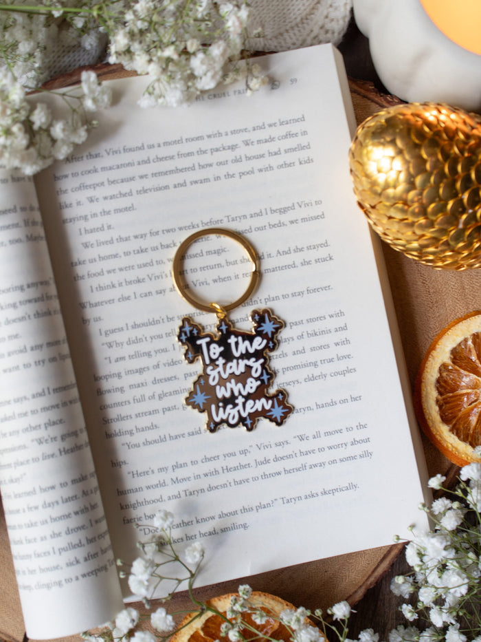 This officially licensed "To The Stars Who Listen" metal keychain is inspired from A Court of Thorns and Roses by Sarah J Maas. It is gold-colored, with the "To The Stars Who Listen" in a white bold script and little light blue stars around it. 